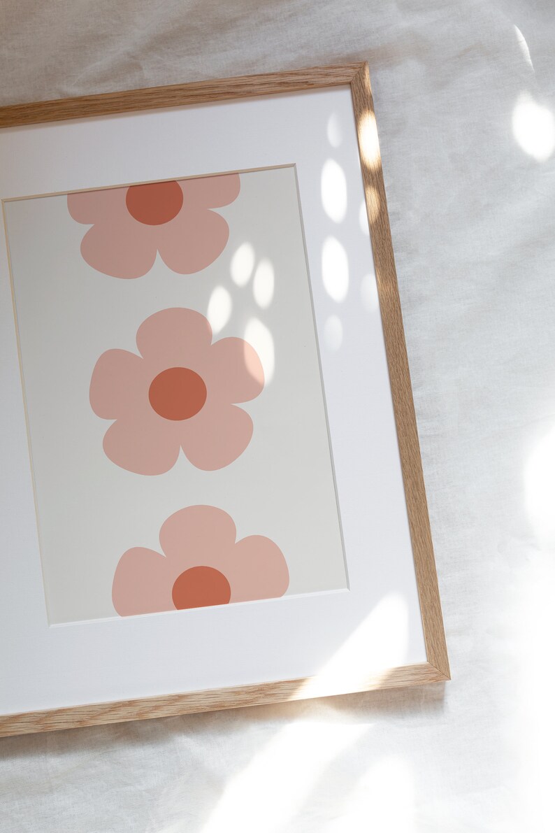 K&ouml;nnte beinhalten: Gerahmter Kunstdruck mit drei stilisierten rosa Blumen mit rostfarbenen Zentren auf cremefarbenem Hintergrund. Das Kunstwerk ist in einem wei&szlig;en Passepartout und einem Naturholzrahmen eingefasst, was eine klare, moderne &Auml;sthetik schafft.