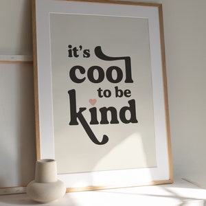 May include: A framed print with the text "it's cool to be kind" in black lettering with a small pink heart above the word "kind". The print is on a cream background and is framed in a light wood frame.