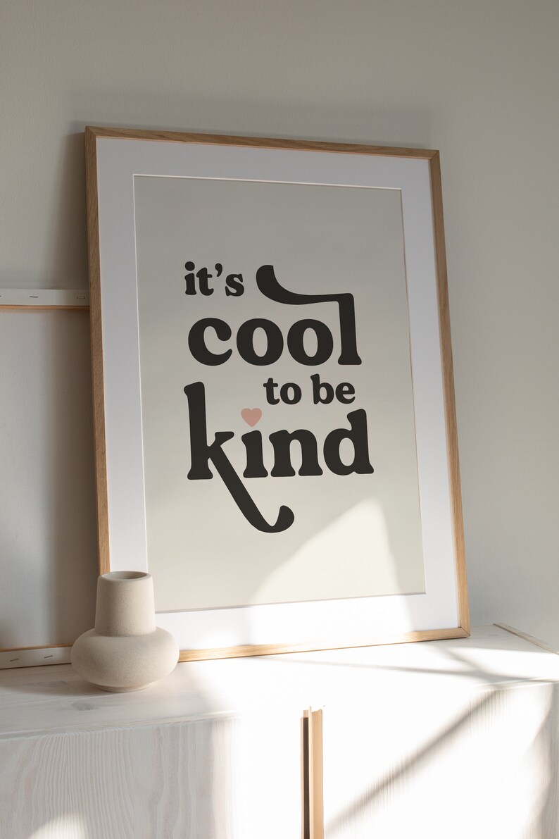 May include: A framed print with the text "it's cool to be kind" in black lettering with a small pink heart above the word "kind". The print is on a cream background and is framed in a light wood frame.