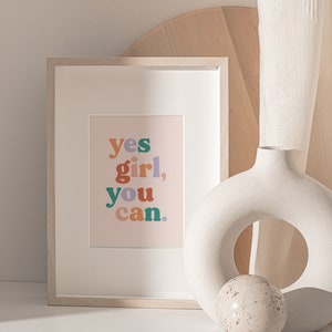 May include: A framed print with the text "yes girl, you can." in colorful letters on a light pink background. The print is sitting on a white surface next to a white ceramic vase with a round opening and a small white sphere.