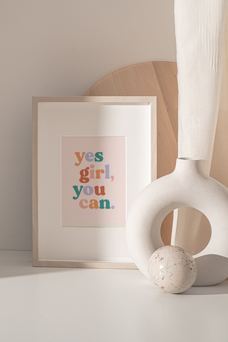 May include: A framed print with the text "yes girl, you can." in colorful letters on a light pink background. The print is sitting on a white surface next to a white ceramic vase with a round opening and a small white sphere.