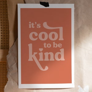 May include: A rectangular art print with a peach background and white border. The print features the phrase "it's cool to be kind" in a retro font. The print is taped to a brown paper surface.