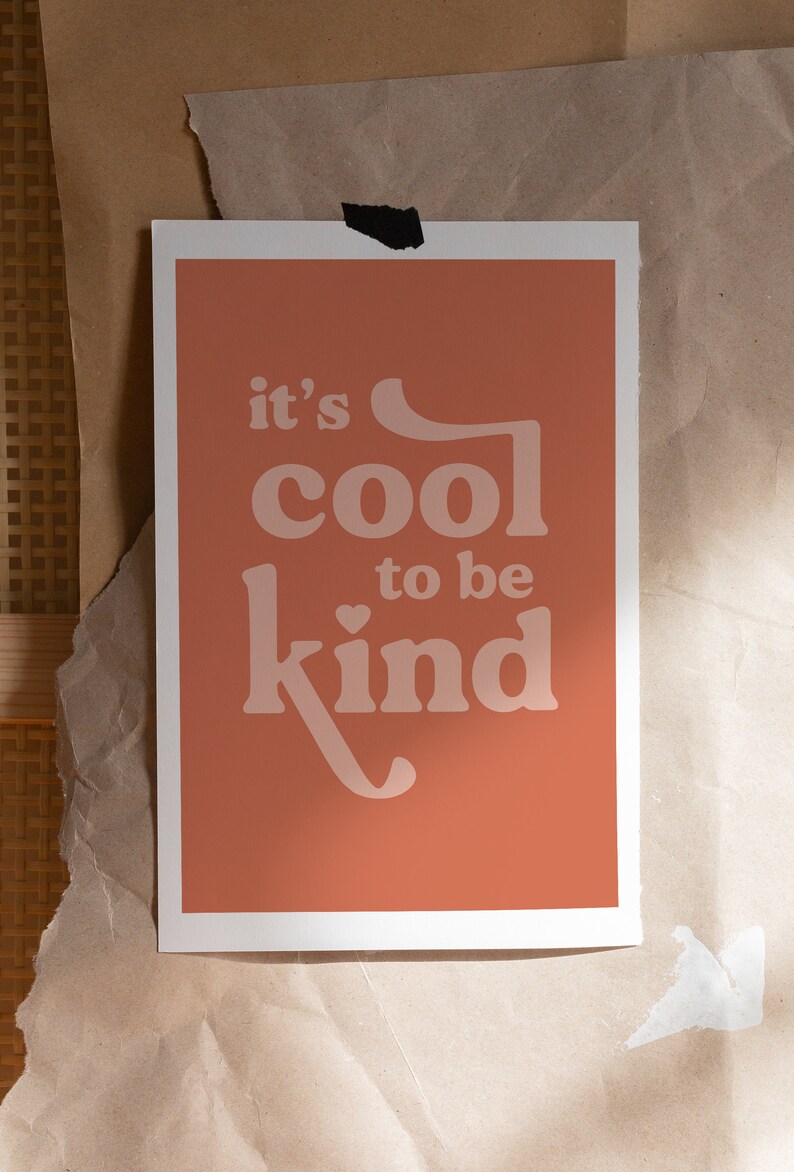 May include: A rectangular art print with a peach background and white border. The print features the phrase "it's cool to be kind" in a retro font. The print is taped to a brown paper surface.