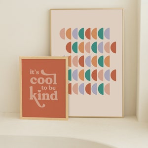 May include: Two framed art prints. The smaller print is a terracotta color with the text "it's cool to be kind" in a retro font. The larger print has a light pink background with rows of colorful half-circle shapes.