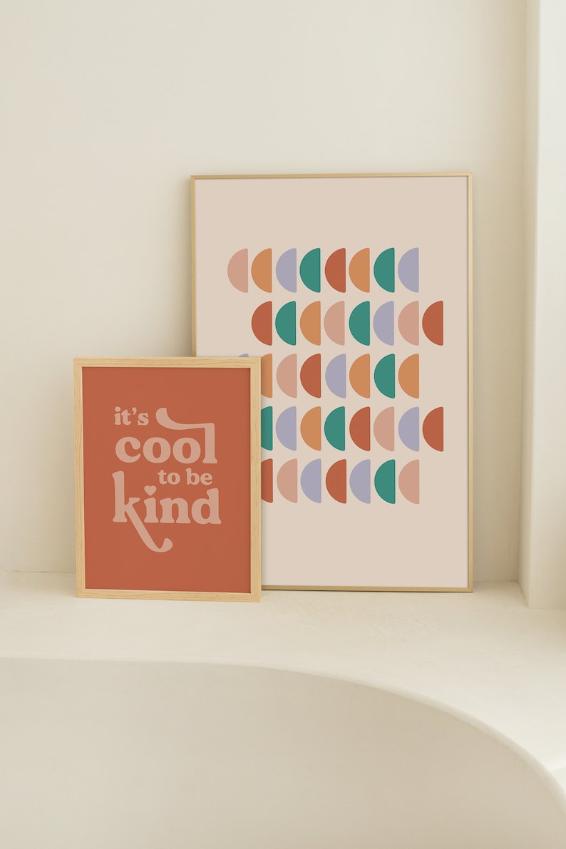 May include: Two framed art prints. The smaller print is a terracotta color with the text "it's cool to be kind" in a retro font. The larger print has a light pink background with rows of colorful half-circle shapes.