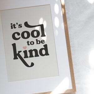 May include: A framed print with the text "it's cool to be kind" in black lettering with a pink heart. The print is framed in a light brown wood frame.