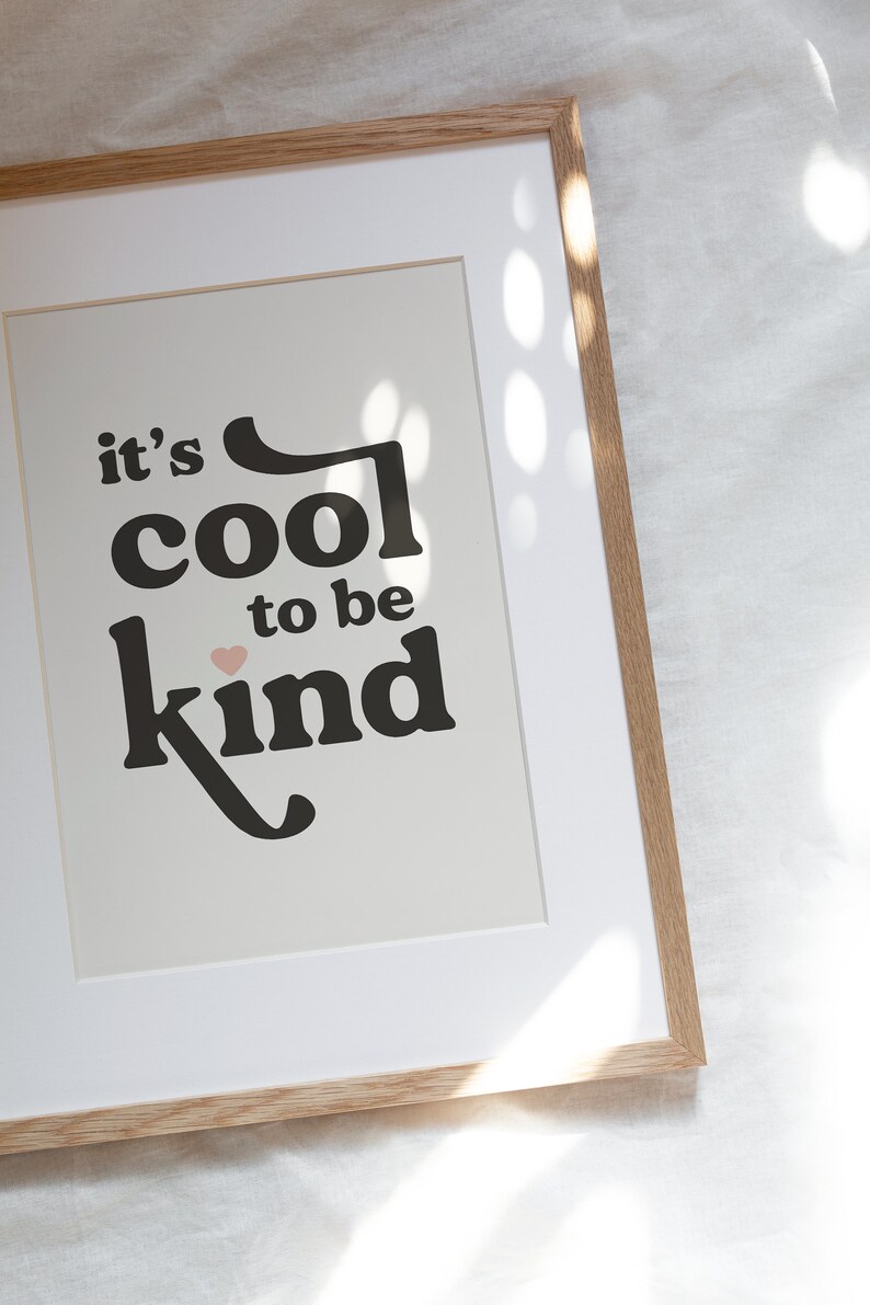 May include: A framed print with the text "it's cool to be kind" in black lettering with a pink heart. The print is framed in a light brown wood frame.