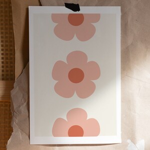 K&ouml;nnte beinhalten: Ein Druck mit drei stilisierten rosa Blumen mit orangefarbenen Zentren auf cremefarbenem Hintergrund. Das florale Design ist auf einem rechteckigen Druck zentriert, der auf einer braunen Papieroberfl&auml;che befestigt ist.