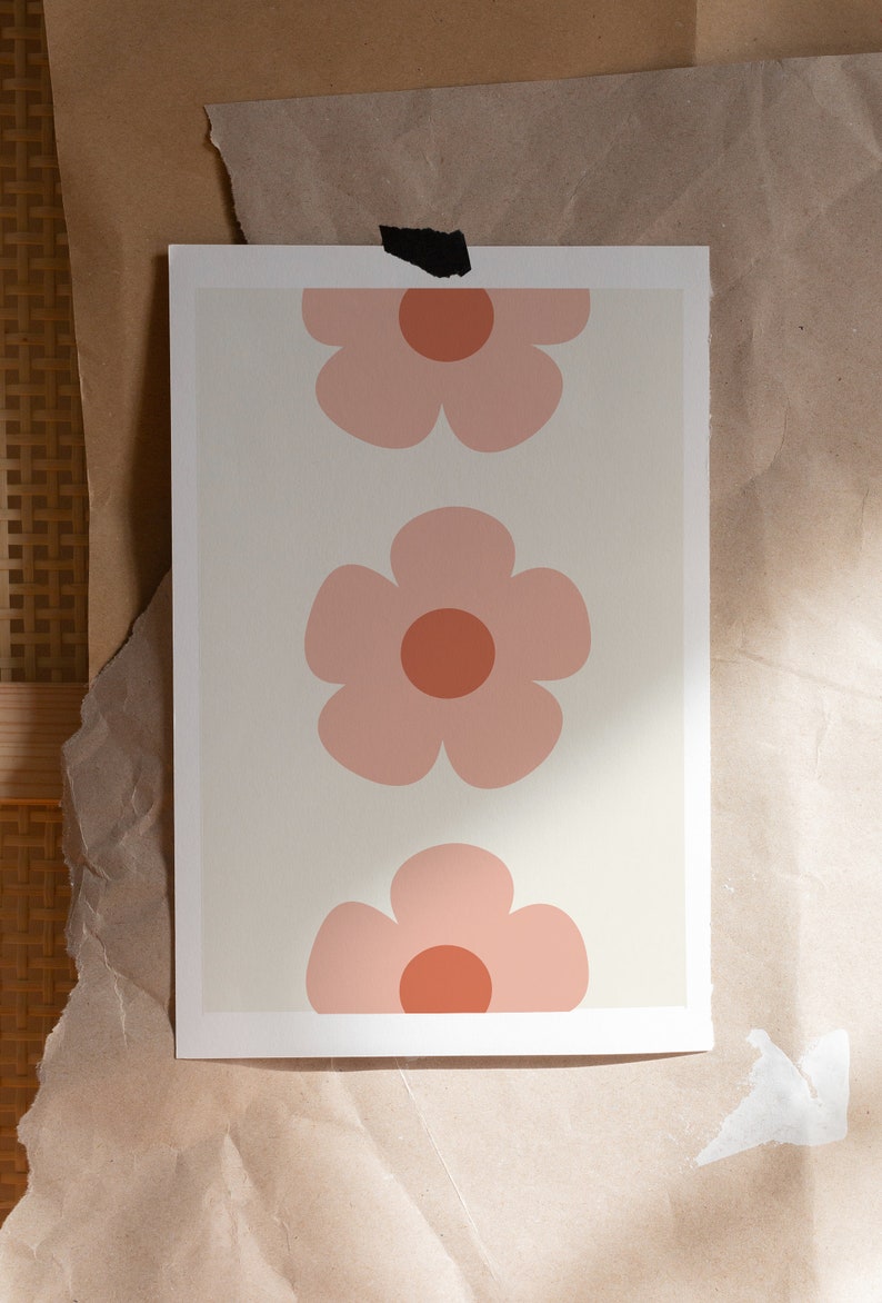 K&ouml;nnte beinhalten: Ein Druck mit drei stilisierten rosa Blumen mit orangefarbenen Zentren auf cremefarbenem Hintergrund. Das florale Design ist auf einem rechteckigen Druck zentriert, der auf einer braunen Papieroberfl&auml;che befestigt ist.
