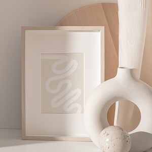 May include: A framed abstract print with a white squiggly line on a beige background, a white ceramic vase with a ring shape, and a small white sphere with brown speckles.
