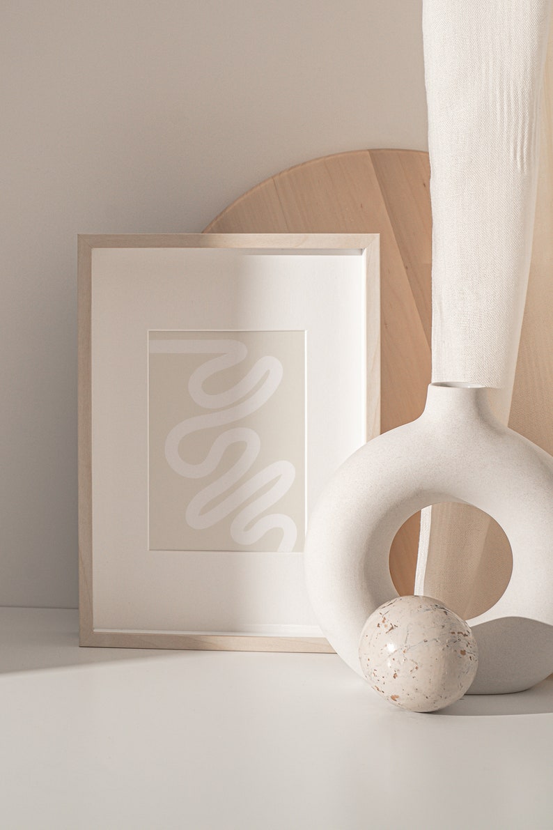 May include: A framed abstract print with a white squiggly line on a beige background, a white ceramic vase with a ring shape, and a small white sphere with brown speckles.