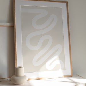 May include: A framed abstract print with a white, wavy line design on a beige background. The frame is made of light wood and the print is sitting on a white surface.