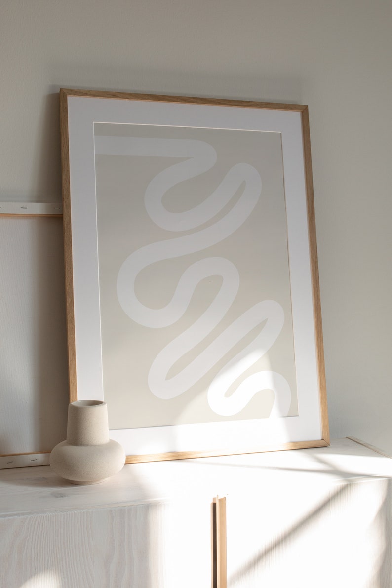 May include: A framed abstract print with a white, wavy line design on a beige background. The frame is made of light wood and the print is sitting on a white surface.