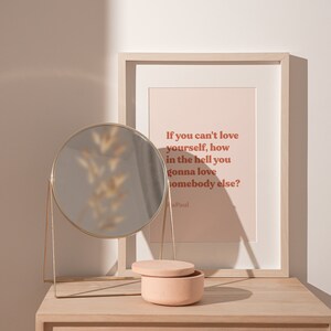 May include: A framed print with the quote "If you can't love yourself, how in the hell you gonna love somebody else?" by Paul. The print is on a light pink background with brown text. The print is on a wooden table with a round gold mirror and a small pink ceramic box.