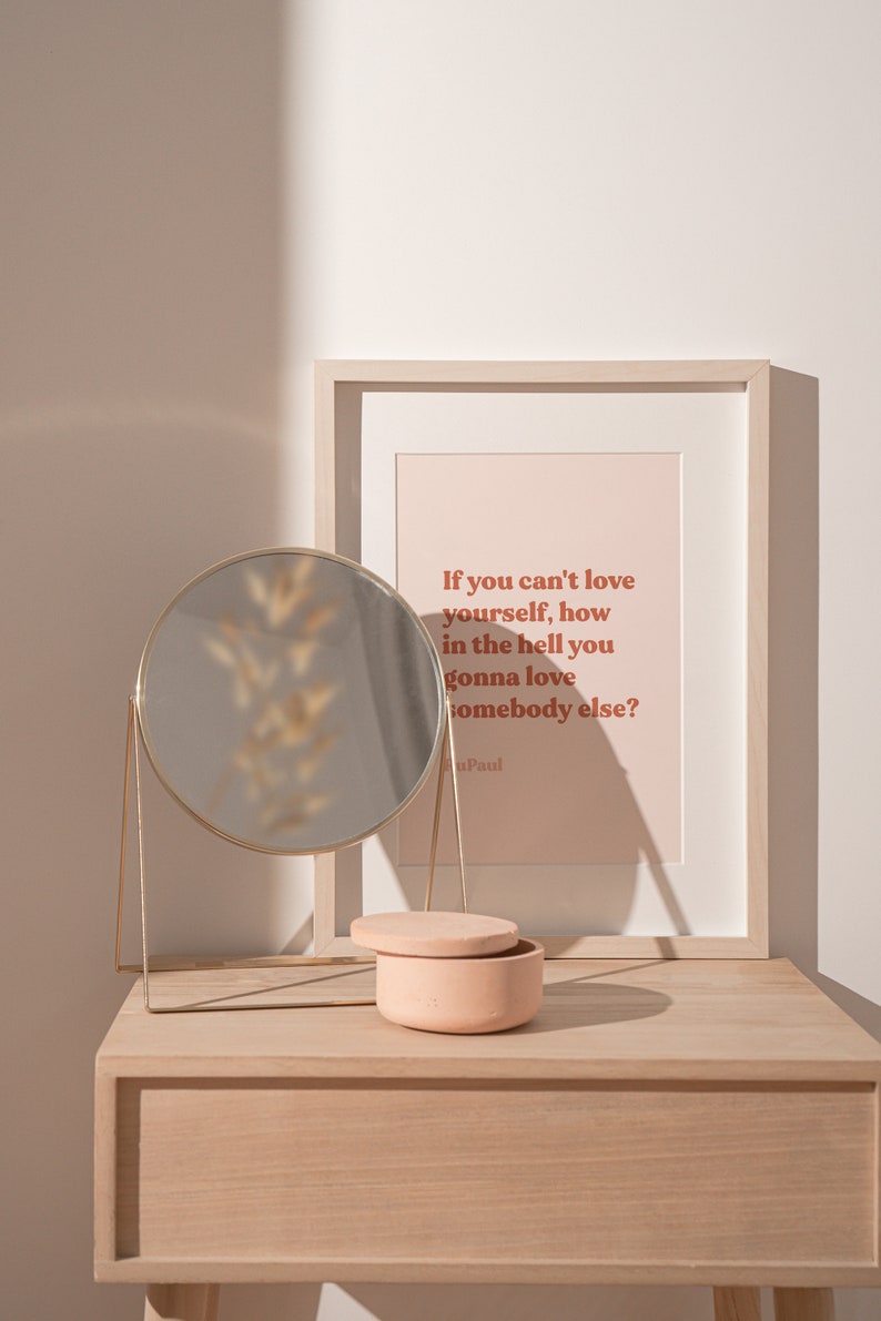 May include: A framed print with the quote "If you can't love yourself, how in the hell you gonna love somebody else?" by Paul. The print is on a light pink background with brown text. The print is on a wooden table with a round gold mirror and a small pink ceramic box.
