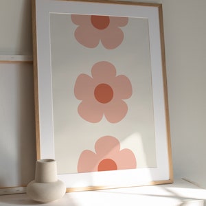 K&ouml;nnte beinhalten: Gerahmter Kunstdruck mit drei gro&szlig;en, stilisierten rosa Blumen mit orangefarbenen Zentren auf cremefarbenem Hintergrund. Das Kunstwerk ist in einem hellen Holzrahmen mit wei&szlig;em Passepartout, geeignet f&uuml;r die Inneneinrichtung.