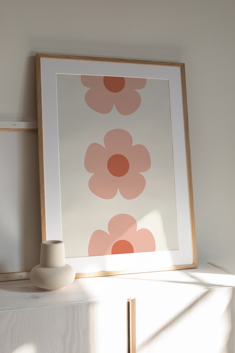K&ouml;nnte beinhalten: Gerahmter Kunstdruck mit drei gro&szlig;en, stilisierten rosa Blumen mit orangefarbenen Zentren auf cremefarbenem Hintergrund. Das Kunstwerk ist in einem hellen Holzrahmen mit wei&szlig;em Passepartout, geeignet f&uuml;r die Inneneinrichtung.