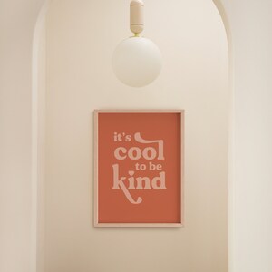 May include: A framed print with the text "it's cool to be kind" in a soft, light pink font against a terracotta background. A wooden stool sits below the print. A white globe light hangs from the ceiling.