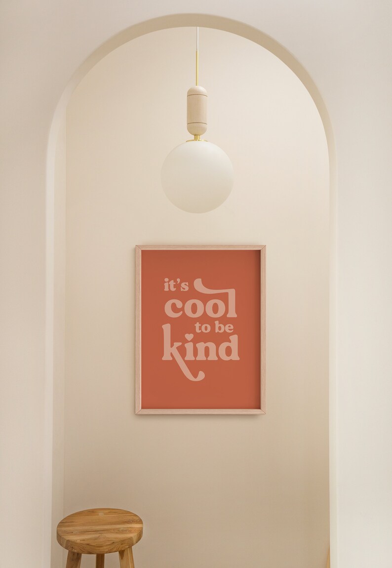 May include: A framed print with the text "it's cool to be kind" in a soft, light pink font against a terracotta background. A wooden stool sits below the print. A white globe light hangs from the ceiling.