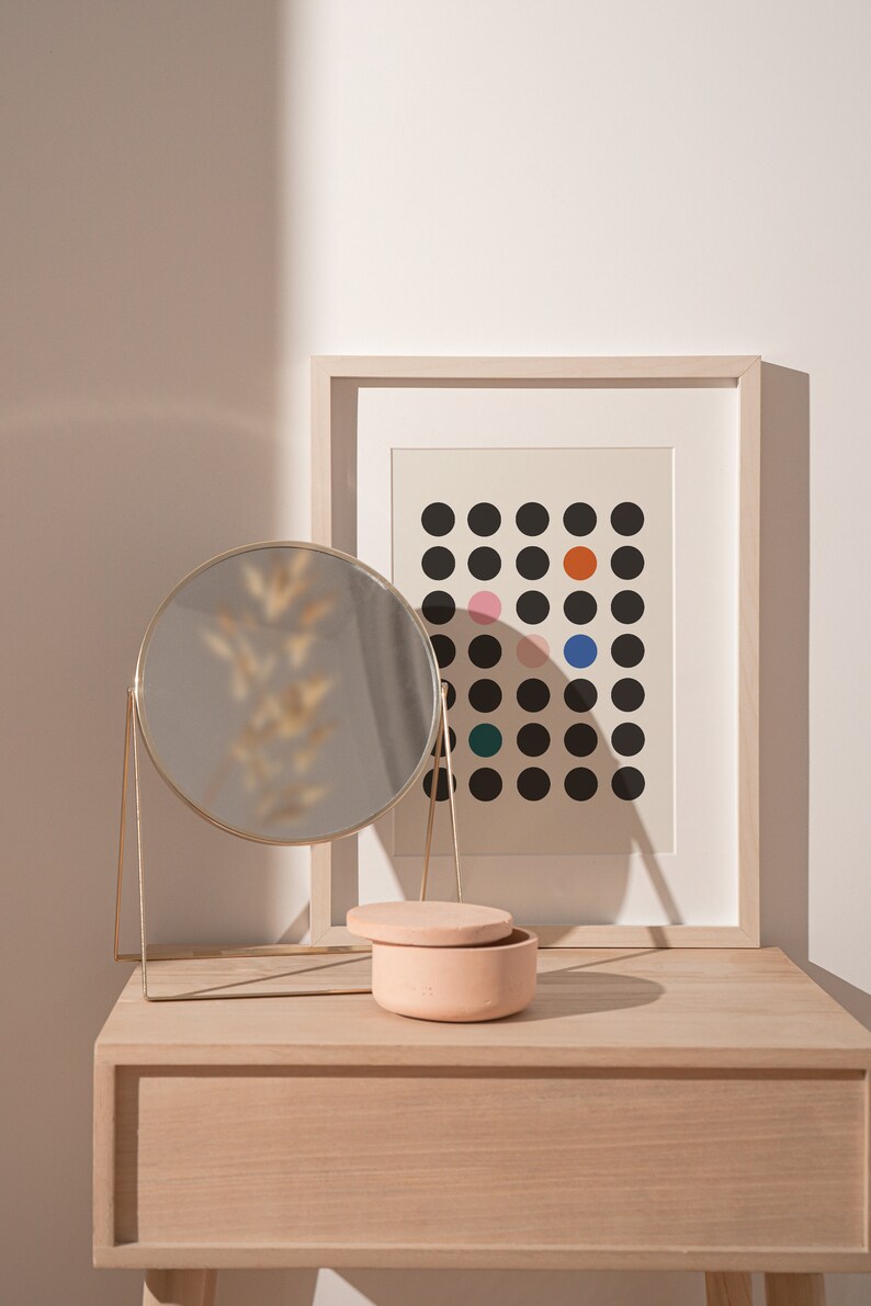 Puede incluir: Una mesa de tocador de madera clara con un espejo redondo dorado, una peque&ntilde;a caja de cer&aacute;mica rosa con tapa y una impresi&oacute;n enmarcada con un patr&oacute;n de lunares negros y blancos con lunares de colores.