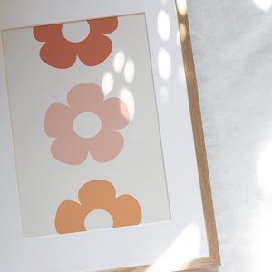 May include: A framed print with a white background and three stylized flowers in shades of orange, pink, and peach. The flowers have a simple, retro design.
