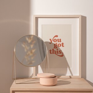 K&ouml;nnte beinhalten: Ein heller Holzschreibtisch mit einem runden goldenen Spiegel, einer kleinen rosafarbenen Keramikdose und einem gerahmten Druck mit dem Text "You got this." in rot-orange.