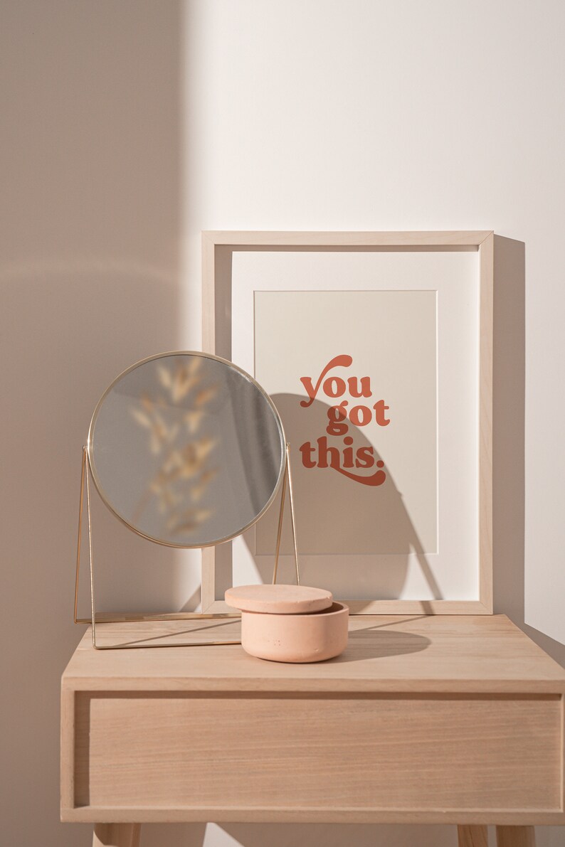 K&ouml;nnte beinhalten: Ein heller Holzschreibtisch mit einem runden goldenen Spiegel, einer kleinen rosafarbenen Keramikdose und einem gerahmten Druck mit dem Text "You got this." in rot-orange.