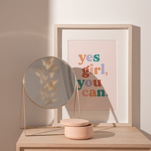 May include: A light wood vanity table with a round gold mirror, a small pink ceramic box, and a framed print with the text "yes girl, you can." in colorful letters.
