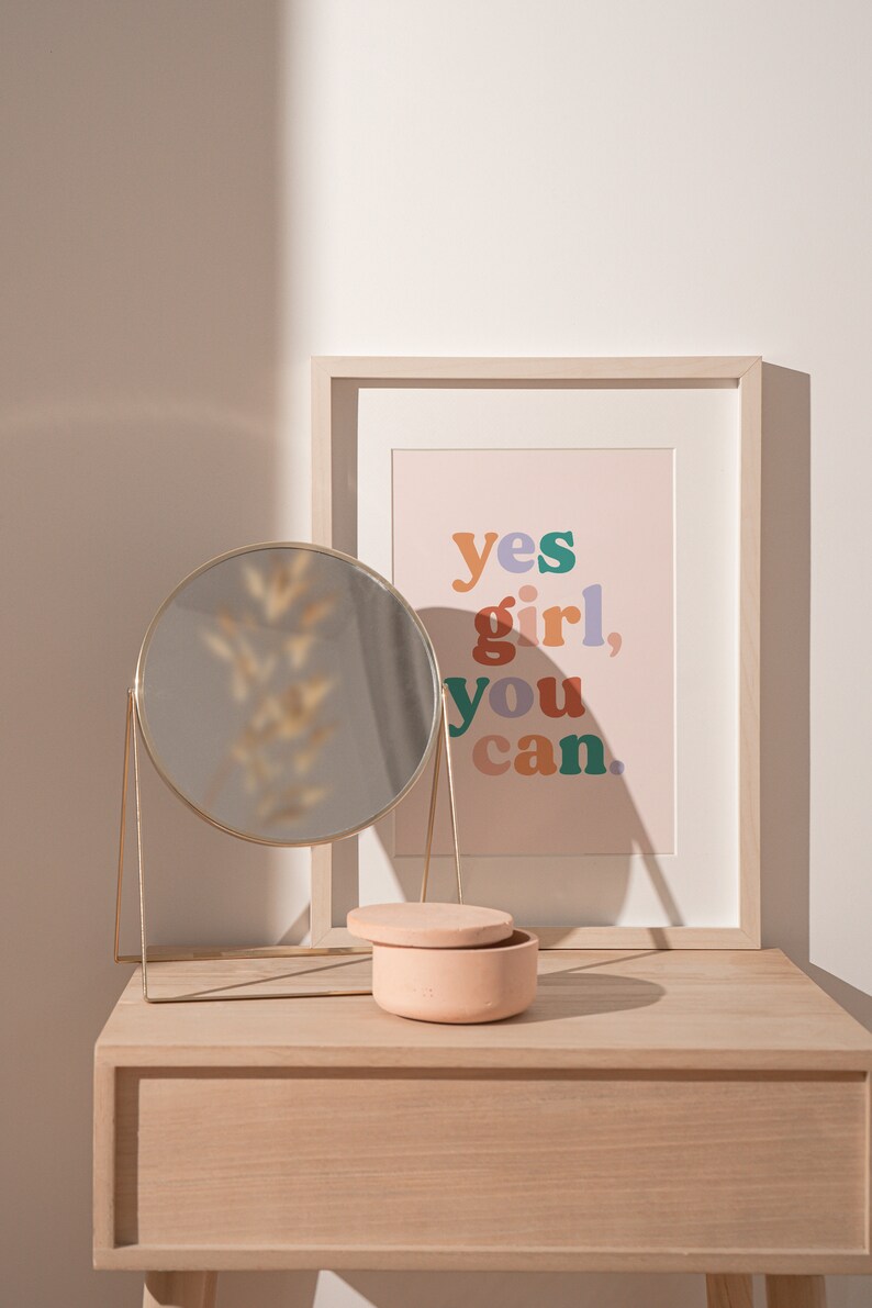 May include: A light wood vanity table with a round gold mirror, a small pink ceramic box, and a framed print with the text "yes girl, you can." in colorful letters.
