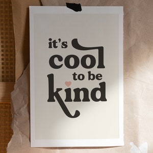 May include: A white framed print with black text that reads "it's cool to be kind" with a small pink heart symbol. The background is a light beige color.