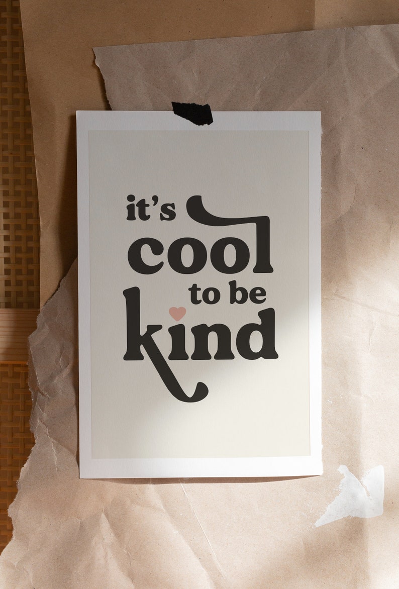May include: A white framed print with black text that reads "it's cool to be kind" with a small pink heart symbol. The background is a light beige color.