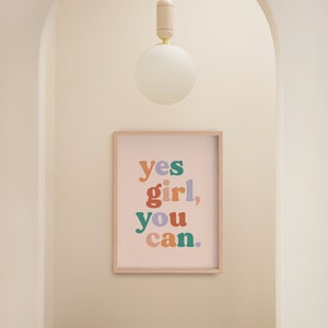 May include: A framed print with the text "yes girl, you can." in colorful letters on a light pink background. The print is hanging on a white wall in a hallway with a wooden stool and a white globe light fixture.