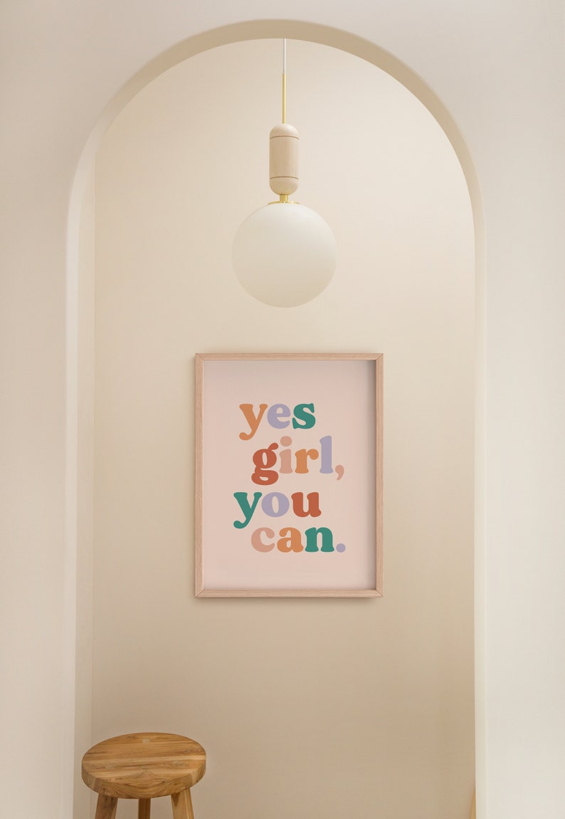 May include: A framed print with the text "yes girl, you can." in colorful letters on a light pink background. The print is hanging on a white wall in a hallway with a wooden stool and a white globe light fixture.