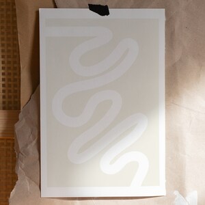 May include: A white abstract line drawing on a light beige background. The lines are curved and flowy, creating a sense of movement. The print is framed with a white border.