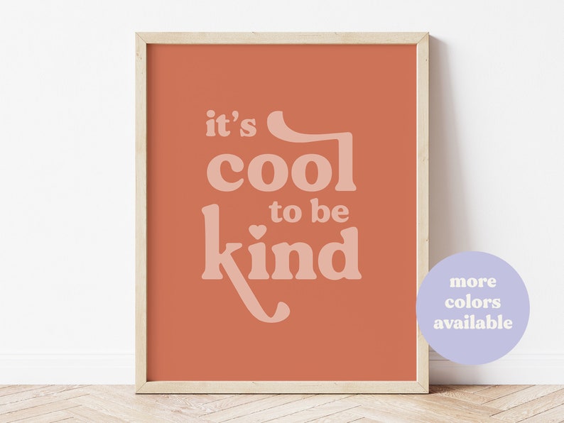 May include: A framed art print with the words "it's cool to be kind" in a light peach color on a terracotta background. The print is in a wooden frame. A small, round, purple graphic in the bottom right corner says "more colors available."