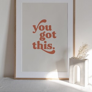 K&ouml;nnte beinhalten: Ein gerahmter Druck mit dem Text "you got this." in einem Retro-Schriftart-Stil. Der Text ist in einem verbrannten Orangeton auf einem hellbeigen Hintergrund. Der Rahmen ist in einer hellen Holzfarbe.