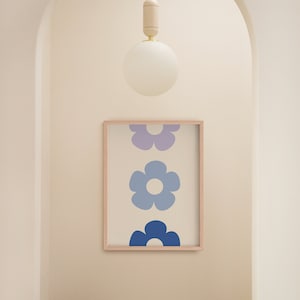 K&ouml;nnte beinhalten: Ein gerahmter Druck mit einer vertikalen Anordnung von drei stilisierten blauen und violetten Blumen. Der Druck h&auml;ngt an einer wei&szlig;en Wand in einem Flur mit einem Holzhocker im Vordergrund. Eine wei&szlig;e Kugel-Leuchte h&auml;ngt von der Decke.