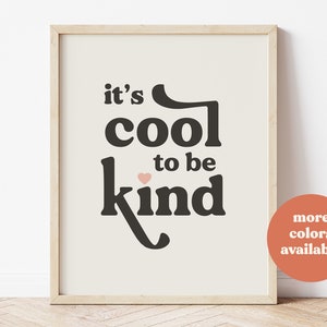 May include: A framed print with the text "it's cool to be kind" in black lettering with a small pink heart above the word "kind". The print has a white background and is framed in a light wood frame.