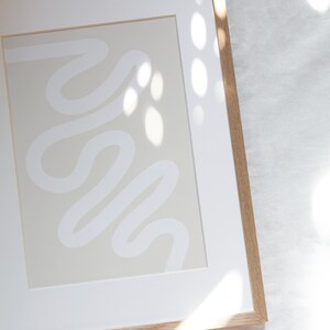 May include: A framed abstract print with a white line design on a light beige background. The frame is made of natural wood.
