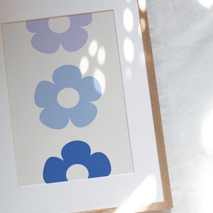K&ouml;nnte beinhalten: Ein gerahmter Druck mit drei stilisierten Blumen in Blaut&ouml;nen und Lila. Die Blumen sind vertikal angeordnet, wobei die gr&ouml;&szlig;te Blume unten und die kleinste Blume oben ist.