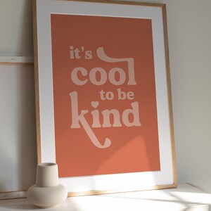 May include: Framed art print with the message "it's cool to be kind" in a soft, retro font. The artwork features a warm, terracotta-colored background and a white mat, encased in a light wood frame. A minimalist vase sits in the foreground.