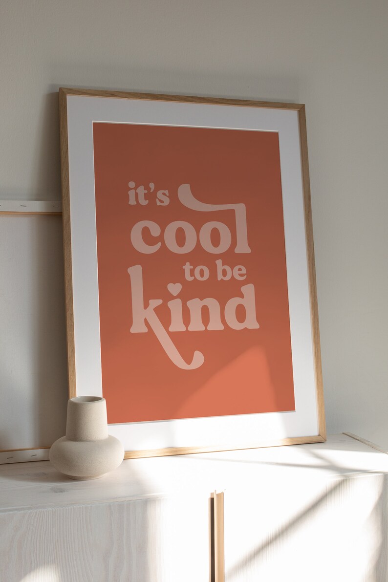 May include: Framed art print with the message "it's cool to be kind" in a soft, retro font. The artwork features a warm, terracotta-colored background and a white mat, encased in a light wood frame. A minimalist vase sits in the foreground.
