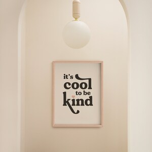 May include: A framed print with the text "it's cool to be kind" in black with a pink heart. The print is hanging on a white wall in a hallway with a wooden stool and a white globe light fixture.