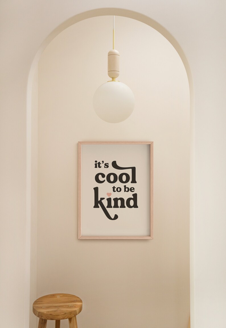 May include: A framed print with the text "it's cool to be kind" in black with a pink heart. The print is hanging on a white wall in a hallway with a wooden stool and a white globe light fixture.