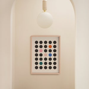 Puede incluir: Una impresi&oacute;n enmarcada de un dise&ntilde;o geom&eacute;trico en blanco y negro con c&iacute;rculos de colores. La impresi&oacute;n est&aacute; colgada en una pared blanca en un pasillo con un taburete de madera y una l&aacute;mpara de globo blanco.