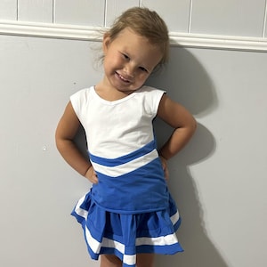 May include: A sleeveless dress featuring a white bodice and a blue skirt section with a chevron pattern. The skirt has a pleated design with horizontal white stripes along the hemline. The garment is constructed with a combination of white and blue fabric.