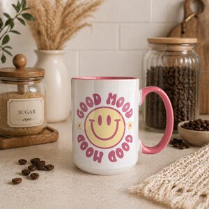 Könnte beinhalten: Weißer Keramikbecher mit rosa Griff und Innenraum. Der Becher zeigt ein gelbes Smiley-Gesicht und die Worte "GOOD MOOD" in Rosa. Der Becher ist von Kaffeebohnen, einem Zuckertopf und dekorativen Gegenständen umgeben.