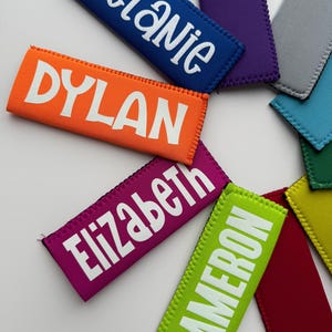 May include: A collection of rectangular neoprene beverage sleeves in various colors including orange, blue, purple, gray, light blue, green, yellow, red, and lime green. Several sleeves feature white printed names: Melanie, Dylan, Elizabeth, and Cameron.