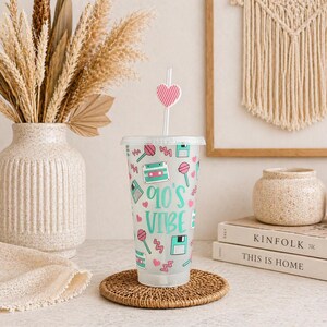 May include: A clear plastic tumbler with a lid and straw, decorated with 90s-themed graphics. The design includes cassette tapes, floppy disks, and lollipops in teal and pink. The text "90's Vibe" is printed on the front. The straw has a pink heart accent.