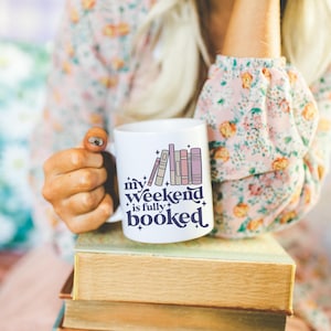 May include: White ceramic mug with the text "my weekend is fully booked" and a graphic of stacked books in shades of purple and beige. The mug is held over a stack of old books.